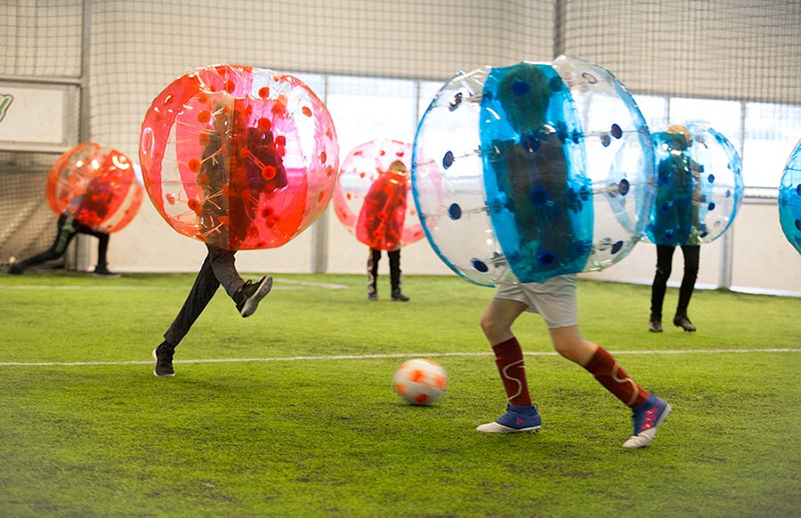 Barn spiller fotball. De har baller i forskjellige farger på overkroppen. Et lag har røde baller og et lag har blå baller på seg. Banen er innendørs og har kunstgress. 